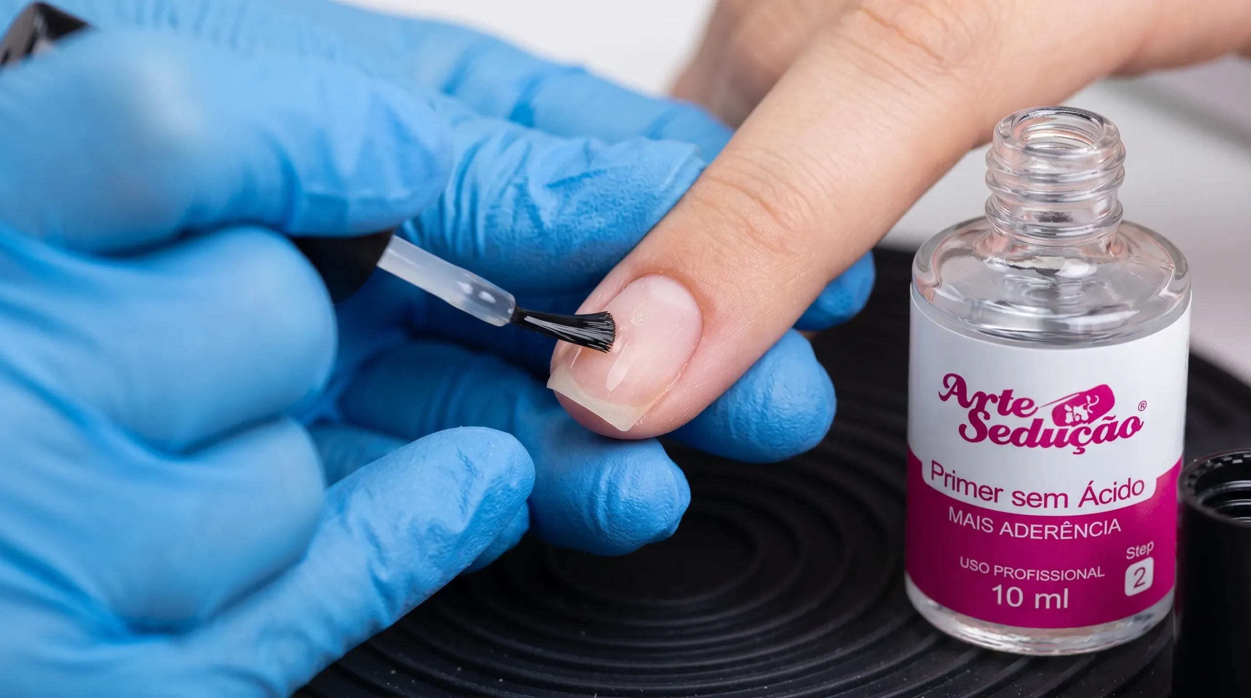 O que vem primeiro, o Primer ou o desidratador? 1 Manicure com luvas aplicando primer transparente em uma unha natural ressecada.